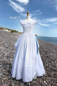 A crisp white cotton dress, full length with a nipped in waist and circle skirt , is shown on a dressmakers dummy on a sunny pebble beach. Blue ribbons flutter at the waist