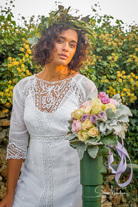 a model with dark, curly hair wears an antique lace wedding gown from the Edwardian era. A crown of laurels and pastel rose bouquet complete the look. 