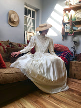A romantic edwardian wedding dress with a full skirt, high neck and lace sleeves - the model is seated on a sofa in her ivory satin gown which is a historical costume replica Edwardian dress with long leg-o-mutton sleeves and guipure lace details 