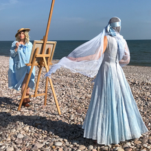 The back view of the pale blue flocked organza original vintage dress - worn by a model whose pale blue antique lace veil is blowing in the breeze. the model stands on a pebble beach. in the background, a second model stands at an easel in a sky blue swing coat from the same era 