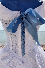 A crisp white broderie anglais dress is laced up the back with a blue satin ribbon, corset style. Around the nipped in waist, you can just glimpse a pebble beach and the sea beyond. 