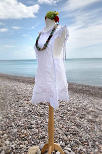 A white linen vintage pinafore dress is shown on a pebble beach, worn with a Boho flowercrown and folky accessories