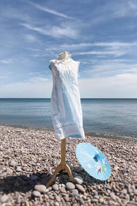 a simple white lawn mini dress with a high neck and drawn thread lace is shown worn on a pebble beach. A parasol is resting on the ground.