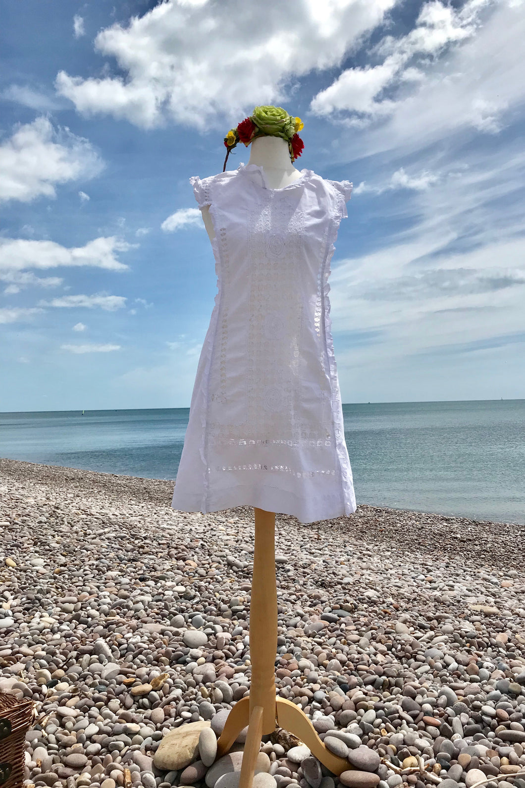 A crisp white linen mini dress and colourful flower crown are shown on a shop dummy with a pebble beach in the background.
