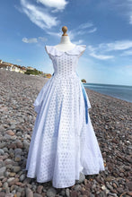 A crisp white cotton dress, full length with a nipped in waist and circle skirt , is shown on a dressmakers dummy on a sunny pebble beach. Blue ribbons flutter at the waist