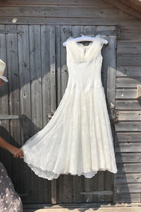 A 50's prom dress in ivory is shown hanging on a beach hut door. A woman is holding the skirt out to show the fullness. 