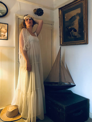 A model is shown in a vintage interior wearing a floor length 1970's flowing white dress in a grecian style with fringed sleeves. The dress has a high waist and three tiers. She also wears a white paper flower crown 