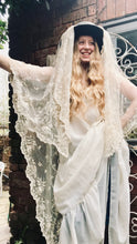 A model with long blonde wavy hair wears a dark hat with a floor length antique lace veil draped over it. She also wears a 1970's flowing wedding dress