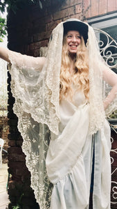 A model with long blonde wavy hair wears a dark hat with a floor length antique lace veil draped over it. She also wears a 1970's flowing wedding dress
