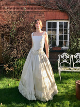 A model wears and antique wedding dress which is in fact two separates - a corset top and full skirt. Both are made from ivory silk. Sunlight dapples the Victorian skirt as the bride stands outside a red brick house.