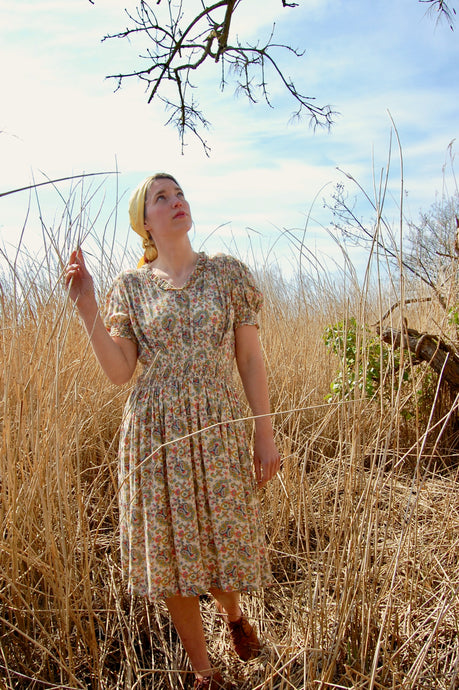 Land girl style! Our model Josie wears a 1940's paisley print summer dress featuring a paisley print, and stands in a field of long, bleached grasses. She has teamed the vintage dress with a vintage silk headdress and flat brown boots. 