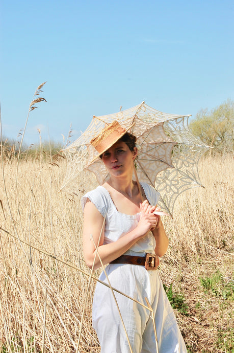 Our model Josie wears an all in one Victorian bloomer set or camobocker in crisp white cotton with a straw hat and parasol. She stands in a field of shoulder high bleached grasses and reeds