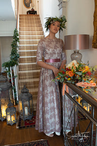 A pretty dark haired model stands in the hallway of a country house, wearing a floor sweeping vintage gown from the 1950's made with a rose patterned lace in an old rose colour. 