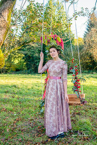 A pretty scene - A dark haired model in a beautiful fifties vintage dress and green leaf crown stands in front of a swing, it's strings twined with flowers. The floor sweeping dress is made of a rose patterned lace in an old rose colour with a deep pink sash. Prom dress wedding inspiration for a bohemian wedding , 
