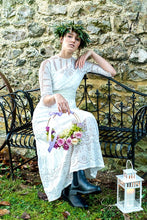 An elegant young woman in a crown of greenery sits on a wrought iron bench. She wears an Edwardian lace wedding dress with broderie and lace panels, a high neck and  elbow length sleeves. She carries a hoop of lilac roses and wears dark boots. 