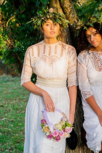 Two young women wear white Edwardian dresses and stand beneath a tree in a ray of golden autumnal sunshine. The central girl wears an original Edwardian white lace wedding dress with broderie anglais panels and hand embroidered details.   