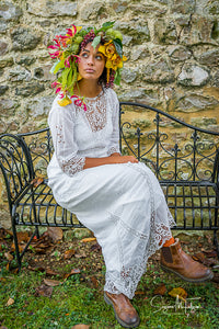 A pretty dark skinned model wears an oversized flower crown in sunset shades of yellow and pink. She is dressed in an antique  Edwardian lace wedding dress with brown leather boots, and is every inch the bohemian bride 