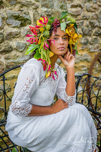 A pretty dark skinned model wears an oversized flower crown in sunset shades of yellow and pink. She is dressed in an Edwardian lace wedding dress. She looks every inch the beautiful boho bride 