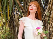 A beautiful willowy red head wears a cream silk 1920's style wedding dress. She stands against a palm tree, with a bouquet of peonies in her hand. 