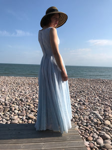 A young girl in a broad brimmed straw hat is backlit on a pebble beach. She wears a vintage fifties dress from Days of Grace in Budleigh Salterton. 