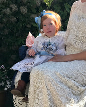 A redheaded toddler girl wears an edwardian bridesmaids dress of white lace 