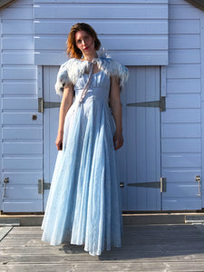A model stands in front of a pale blue beach hut wearing a pale blue organza gown from the 1950's, flocked with a floral pattern. Around her shoulders is a sky blue feather cape. She is sun dappled, and the breeze ruffles her skirts a little 