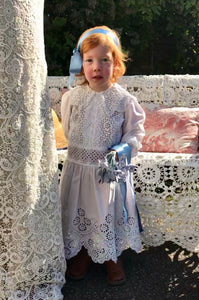 Alice in Wonderland style! A red haired toddler stands in an Edwardian bridesmaids dress made of crisp white cotton lace. She holds a bunch of blue silk flowers and has a blue satin alice band