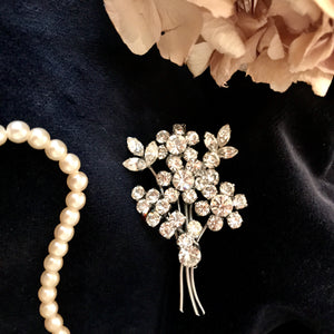 a spray of flowers formed from glittering white diamantes are mounted on a pin brooch - the original fifties brooch is sat on dark velvet, surrounded by a string of pearls and some flower petals 