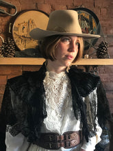 a beautiful prairie girl wears an Edwardian replica wedding dress in ivory satin and lace with a high lace neck. The dress is accessorised with an antique shawl of black lace, a battered cowboy hat and a brown leather belt. The prairie cowgirl bride is lit by a beam of warm sunlight, and stands in front of a rustic fireplace.  