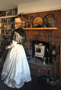 Back view - our prairie cowgirl bride stands in front of an old woodburner in a rustic boho room. Her floor sweeping ivory satin Edwardian style wedding dress is teamed with a cowgirl hat, antique black lace mantilla and brown leather waist cincher belt  