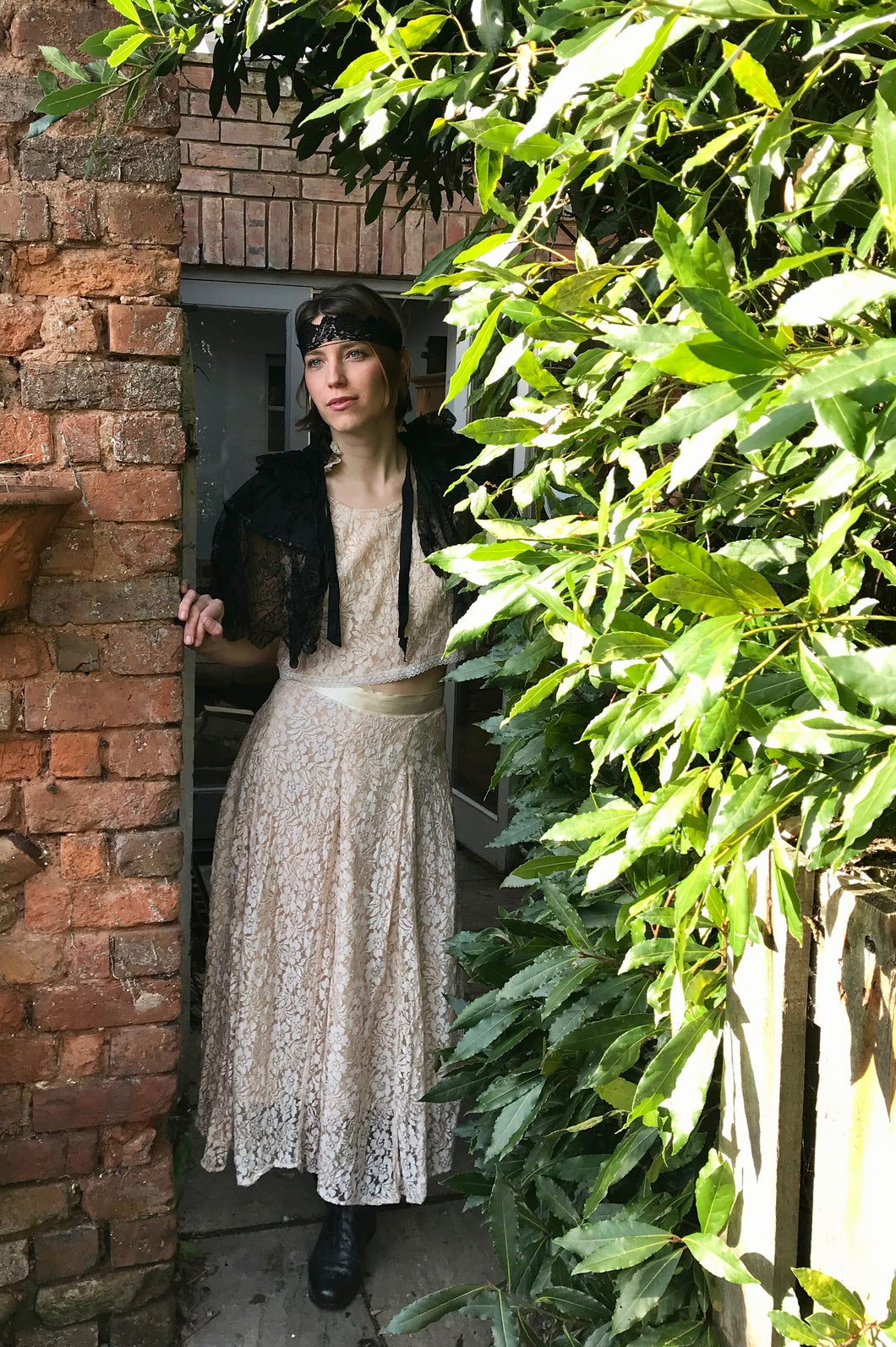 A pretty girl peers through a garden doorway. She is wearing 1940's separates made in shell pink lace. An antique black lace headband and cape complete the Bloomsbury style look. 