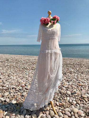 An Edwardian lace wedding dress with antique Honiton lace motifs, a dropped waist and elbow length sleeves is shown on pebble beach with floral crown of pink silk peonies 