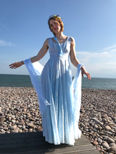 A girl in an elegant sky blue gown from the fifties is backlit by bright sunlight. She stands on a pebble beach, a soft blue veil blows in the breeze