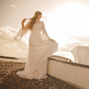 romantic seventies wedding dress with angel sleeves - a blonde model wears an iconic 1970's wedding dress shown backlit by stormy light on Sidmouth beach in Devon 