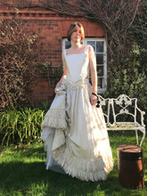 An elegant model stands in front of a red brick cottage, wearing a Victorian vintage ivory silk corset and skirt. She holds a gathered handful of the lace adorned skirt, showing it's fullness. A hat box sits on the grass beside her, and her expression is wistful. Worn together, the corset and skirt appear as one gown, perfect for a wedding dress.