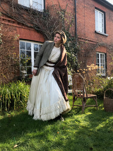 A whimsical bride wears her Victorian wedding dress with a 1930's tweed jacket, arran knit jumper, antique paisley shawl and double brown leather belt. She stands outside a red brick cottage with the sunlight streaming across her