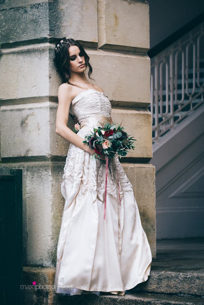A dark haired model in a vintage gown leans against the wall of a historic building. She wears an oyster satin 1950's strapless dress with a ruched bodice and folded collar. She carries a bouquet of cream, pink and deep red flowers